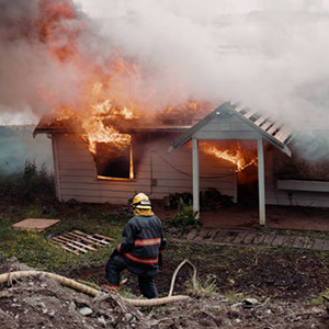 A woman firefighter works to move equipment, a burning unrecognizable home fully engulfed in fire and smoke in the background.